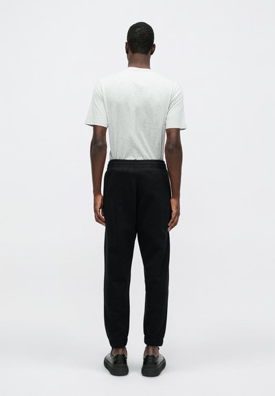 Tricou scurt, de culoare gri deschis, cu mâneci scurte, purtat cu pantaloni de trening negri, cu talie elastică și manșete ajustate; modelul este întors cu spatele.