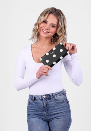 Black wallet with large white polka dots, featuring a gold accent. A model holds it while wearing a white top and blue jeans.