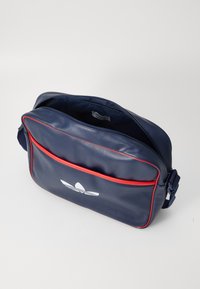 Bolsa de ombro em couro azul-marinho com detalhes em vermelho, apresentando um fecho de zíper e um bolso frontal com um logótipo branco. Textura suave e alça almofadada.