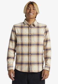 Camisa de franela con botones en un patrón de cuadros beige y púrpura, con cuello, mangas largas y un bolsillo en el pecho. Textura suave.
