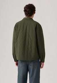 Veste matelassée vert olive avec un col et des poignets boutonnés, portée avec un jean en denim bleu, vue de dos. Tissu texturé avec des lignes verticales.