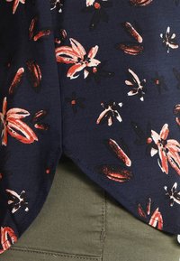 Námořnická květinová blůza s červenými a bílými květinovými vzory, měkkou texturou a zakřiveným spodním lemem, sladěná s olivově zelenými kalhoty.