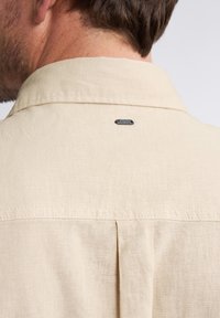 Lichtbeige shirt van een gestructureerde stof, met een rugplooien en een klein zwart metalen logo-accent bij de kraag.