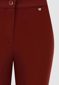 Pantaloni bordeaux con una texture liscia, chiusura frontale con bottoni e un dettaglio in oro. Progettati per una silhouette aderente.