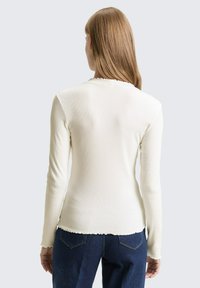 Vrouw met recht, lichtbruin haar die een aangesloten witte top met lange mouwen en donkerblauwe spijkerbroek draagt, gezien van achteren tegen een effen achtergrond.