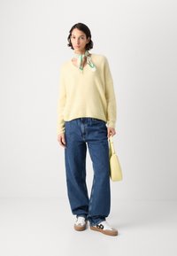 Gelber Strickpullover mit V-Ausschnitt, blaue weite Jeans, weiße Sneakers mit beigen Akzenten und eine kleine gelbe Handtasche. Grüner gemusterter Schal um den Hals gebunden.