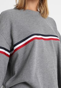 Harmaa ribbimekko, jossa on pyöreä kaula-aukko, ja rinnan kohdalla vaakasuoria punaisia, valkoisia ja tummansinisiä raitoja.