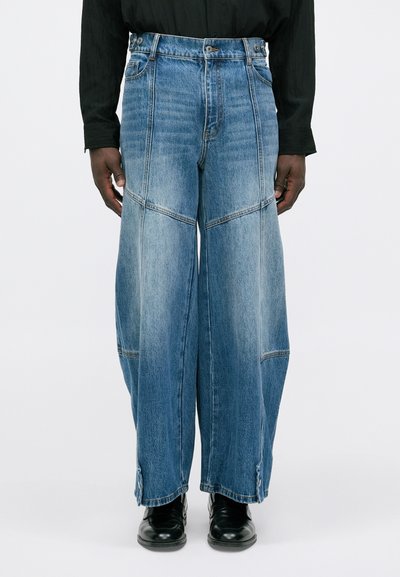 Wide-leg blue jeans with panel stitching, front pockets, and front and back waistband worn with black shoes and a black long-sleeve top.