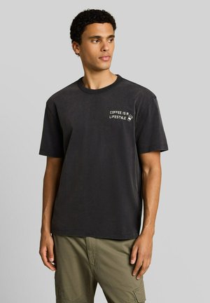 Mann trägt schwarzes T-Shirt mit "Coffee is a lifestyle" Text und Grafik, kombiniert mit olivgrüner Cargohose, schaut nach links.