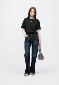 Μαύρο φαρδύ T-shirt με λογότυπο, συνδυασμένο με σκούρα μπλε τζιν καμπάνα. Το μοντέλο κρατά μια πράσινη τσάντα με σχέδια και φορά σαγιονάρες με λουράκια.