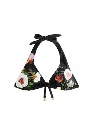 Žena si upravuje vlasy, oblečená v čiernych kvetovaných bikinách, stojaca na skalnatom pobreží s oceánskymi vlnami v pozadí.