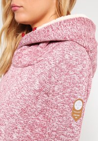 Punainen ja valkoinen melange-huppari, jossa on pehmeä fleecevuori. Hupparissa on suuri huppu, pitkät hihat ja pyöreä logo-piiri hihassa.