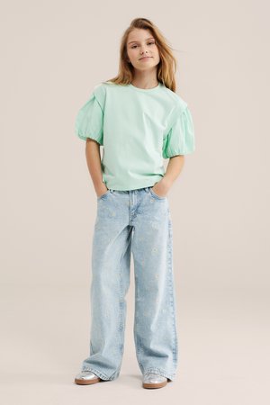 Mlada deklica stoji z rokami v žepih, oblečena v mintasto zeleno majico s nabranimi rokavi, široke svetlomodre kavbojke s cvetličnim vzorcem in srebrnimi čevlji.