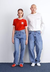 Duas pessoas estão lado a lado. Uma veste uma t-shirt vermelha da Adidas e uns jeans largos de lavagem clara. A outra veste uma camisa de mangas compridas branca e jeans semelhantes.