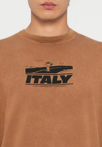 Brun bomull T-skjorte med rund hals. Har et svart grafisk design som viser en tegneseriefigur og teksten "ITALIA".