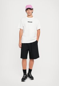 Hvit t-skjorte med korte ermer og svart logo, sammen med svarte shorts. En rosa caps og sorte joggesko med hvite detaljer fullfører antrekket.