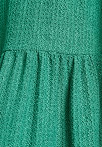 Tecido verde texturizado com um padrão ondulado, apresentando uma linha de cintura franzida que cria uma silhueta evasé. Material suave e leve.