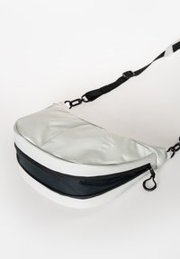 Silberne und schwarze Schultertasche mit einem geschwungenen, gepolsterten Design, ausgestattet mit einem Reißverschlussfach und einem verstellbaren Schultergurt. Strukturierte Oberfläche.