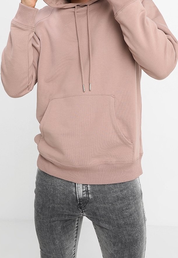Osoba v dusty růžové mikině s kapucí a přední kapsou, v kombinaci se světlými černými slim-fit džíny, na jednolitém pozadí.