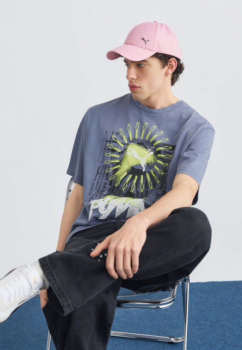 Νεαρός άντρας που φοράει ροζ καπέλο, μπλε μπλουζάκι Puma με σχέδιο, μαύρο παντελόνι και λευκά αθλητικά παπούτσια, καθισμένος σε καρέκλα με σταυρωμένα πόδια.