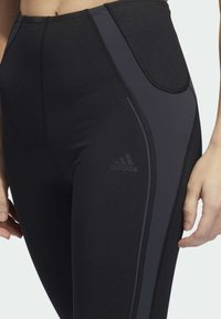 Schwarze Sportleggings mit einem strukturierten hohen Bund, glattem Körper, reflektierendem Seitestreifen und Adidas-Logo auf der unteren linken Seite.