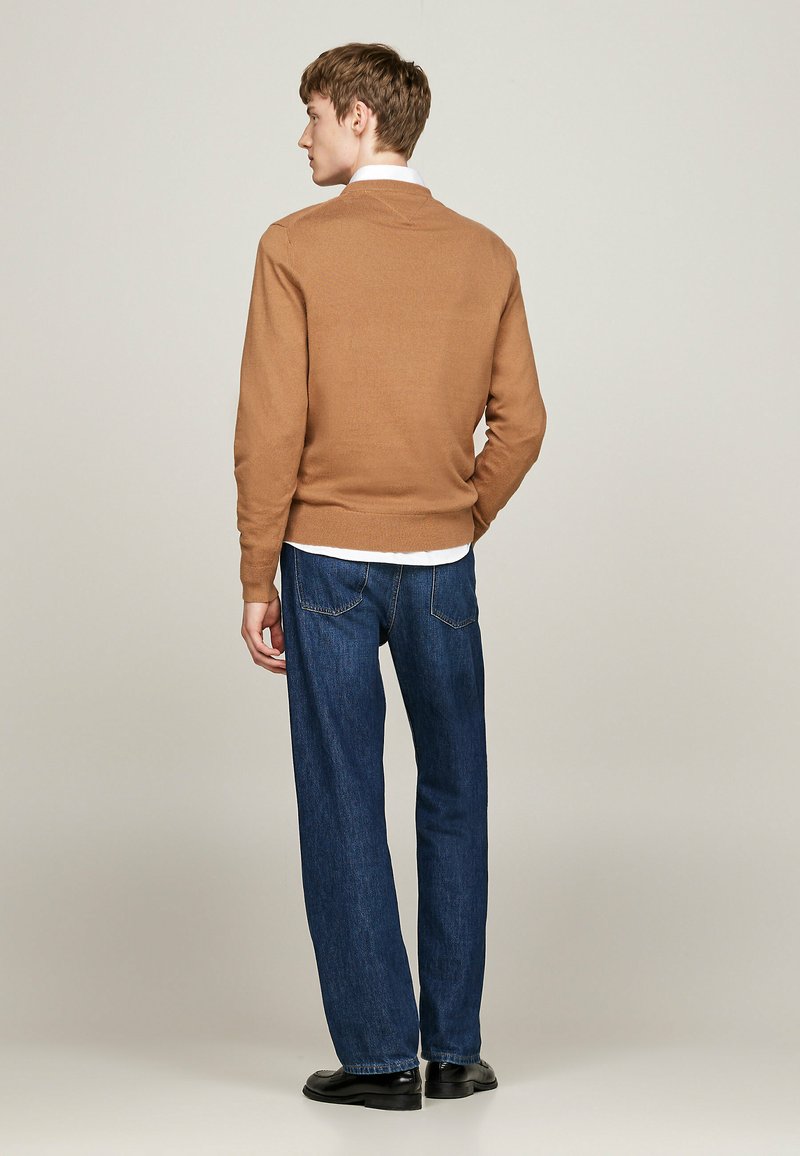 Brauner gestrickter Pullover mit V-Ausschnitt, kombiniert mit blauen Jeans im geraden Schnitt. Glatte Textur, minimale Details, getragen mit schwarzen Schuhen.