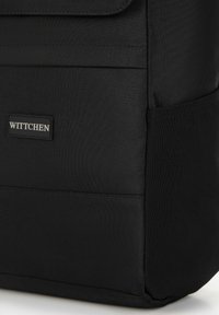 Mochila de tela negra con una textura suave, que presenta un cierre de solapa y una etiqueta de logo visible. Diseño simple y rectangular.