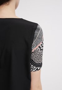 Femme portant un haut noir à manches courtes montrant des motifs géométriques et ondulés noirs et blancs complexes avec une touche de détail rose.