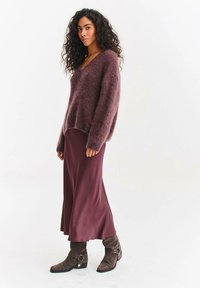 Žena s kudrnatými vlasy, oblečená do načechraného fialového svetru, ladící fialové saténové sukně a hnědých kotníkových bot v neutrálním prostředí.