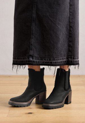 Bottines noires en matériau semblable au daim, avec un talon épais, des panneaux latéraux élastiques et des accents texturés. Portées avec une jupe noire déchirée.