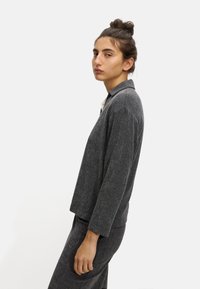 Šedá límcová bunda, texturovaná tkanina, volný střih, boční pohled, spárovaná se shodnými kalhotami, jednoduchý design a neutrální barvy.