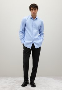 Uomo con camicia azzurra chiara a bottoni, pantaloni neri a righe e scarpe eleganti nere, in piedi con le mani nelle tasche.
