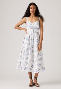 Donna con lunghi capelli ricci che indossa un vestito midi senza maniche bianco con stampe floreali blu e sandali neri con cinghie, in piedi contro uno sfondo neutro.