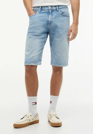 Mand iført lyseblå denimshorts, hvide sokker med logo og beige snørede afslappede sko, stående med den ene hånd i lommen.