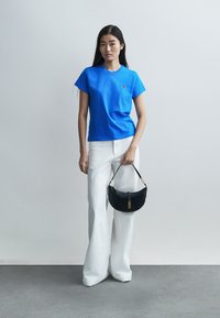 Μπλε βαμβακερό μπλούζα με κοντά μανίκια και μικρό λογότυπο, συνδυασμένο με λευκό παντελόνι φαρδιάς γραμμής και μικρή μαύρη δερμάτινη τσάντα.