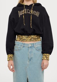 Donna che indossa un hoodie corto nero di Just Cavalli con rifiniture in stampa animale gialla e una gonna di jeans chiara con spacco anteriore.