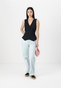 Black sleeveless waistcoat with peplum shape and button detail, paired with light blue jeans, holding a small pink handbag.