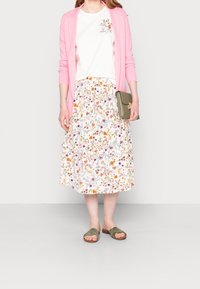 Cárdigan rosa claro sobre una camiseta blanca con bordados florales; falda floral con volantes y sandalias deslizantes en color verde oliva.
