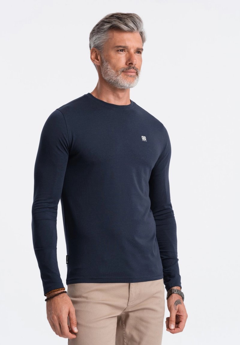 Laivastonsininen pitkähihainen t-paita, joka on valmistettu pehmeästä kankaasta. Paidassa on pyöreä kaula-aukko ja pieni logo rinnassa. Yhdistetty vaaleisiin housuihin.