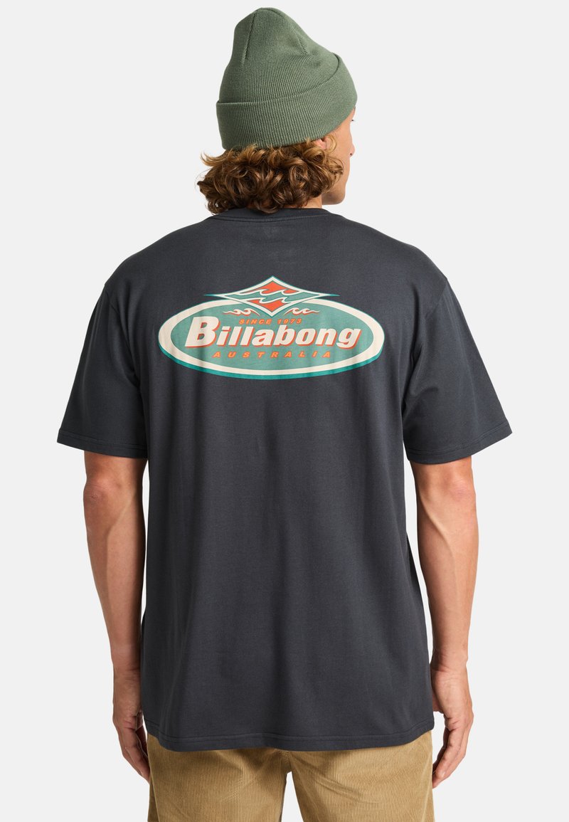 Grijze katoenen t-shirt met een groot, kleurrijk "Billabong" logo op de achterkant. Model draagt een groene beanie en wordt van achteren weergegeven.