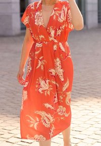 Personne portant une robe midi orange à fleurs avec un profond décolleté en V, debout dans une rue pavée baignée de soleil. Visage non visible.