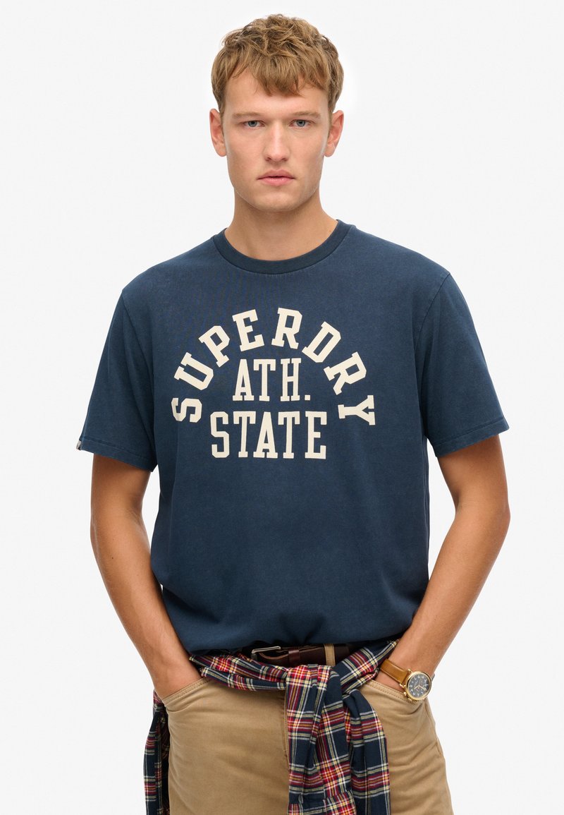 Camiseta de algodón azul marino con un gran gráfico color crema que dice "SUPERDRY ATH. STATE," combinada con pantalones beig y una camisa de cuadros atada a la cintura.