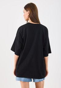 Tricou negru oversized cu mâneci scurte, cu guler rotund și o croială lejeră, purtat împreună cu pantaloni scurți din denim ușor albastru, uzati.