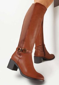 Bottes en cuir marron montantes avec une finition lisse, un talon carré, une fermeture éclair sur le côté et un détail de boucle décoratif à la cheville.