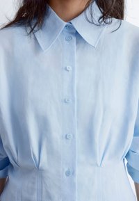 Light blue collared shirt made of soft fabric, featuring six front buttons and subtle pleats at the waist for a structured fit.