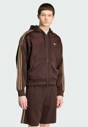 Junger Mann trägt einen braunen Adidas Trainingsanzug mit beigen Streifen, einschließlich eines Zip-Hoodies und passenden Shorts, stehend vor einem einfarbigen Hintergrund.