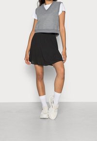 Vrouw in een grijze mouwloze top boven een witte T-shirt, zwarte geplooide rok, witte sokken en stevige witte sneakers, staande tegen een neutrale achtergrond.