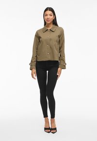 Olive green cropped jacket with a pointed collar, five large buttons, and two side pockets, paired with black leggings and heeled sandals.