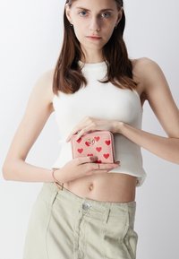 Roze portemonnee met een rode hartprint, gemaakt van textuurmateriaal. Voorzien van een logo en een ritsluiting, vastgehouden door een persoon in een witte top en lichte broek.