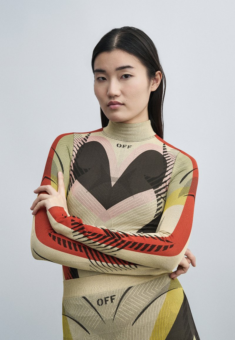 Νέα γυναίκα με ίσια μαύρα μαλλιά, που φοράει ένα μακρυμάνικο τοπ με σχέδια και φούστα με αφηρημένα σχήματα και τη λέξη "OFF", σταυρωμένα χέρια.